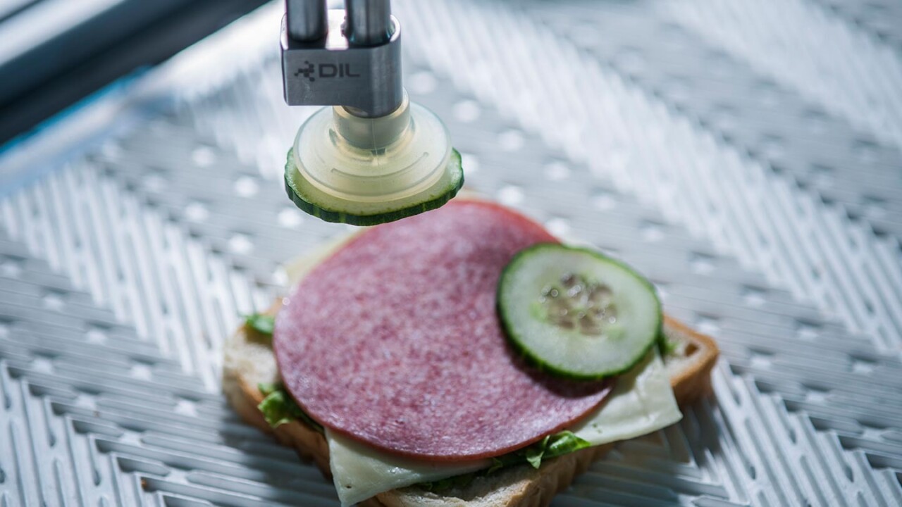 Ein automatisierter Greifer/Roboterarm mit DIL-Logo platziert eine Gurkenscheibe auf ein belegtes Brot (mit Wurst und Käse) auf einer Förder- bzw. Arbeitsfläche. Das Bild zeigt Automatisierung und Robotik in der Lebensmittelproduktion sowie präzise, reproduzierbare Bestückungsprozesse.