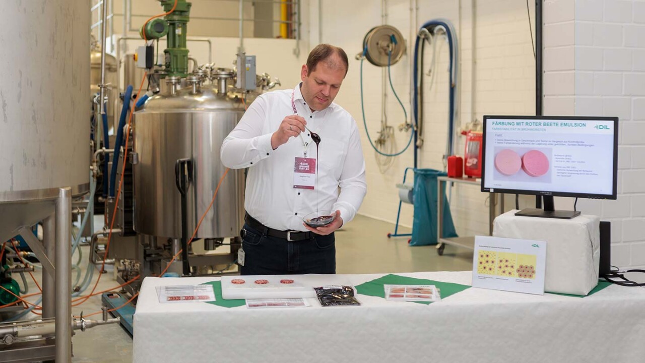 Ein Mitarbeiter demonstriert in einer Pilotanlage die Verarbeitung bzw. Applikation eines Produkts an einem Probentisch. Im Hintergrund sind Edelstahltanks und Anlagentechnik zu sehen; rechts zeigt ein Monitor eine Präsentationsfolie zur „Färbung mit roter Bete Emulsion“ mit Produktabbildungen. Auf dem Tisch liegen Proben und Informationsmaterial.
