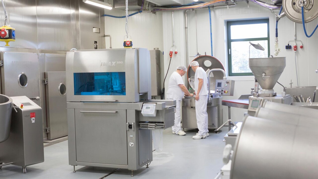 Blick in eine industrielle Pilotanlage mit Edelstahlmaschinen und Prozesslinien. Zwei Mitarbeitende in Hygienekleidung arbeiten an einer Produktionsanlage zur Verarbeitung und Optimierung von Lebensmitteln. Das Bild steht für angewandte Forschung, Prozessentwicklung, Skalierung vom Labor in den Technikum-/Industrie-Maßstab sowie Qualität und Sicherheit in der Lebensmittelproduktion.