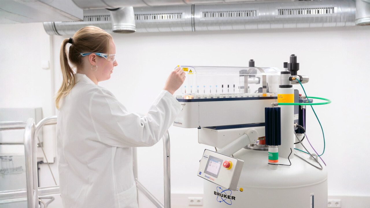 Laboraufnahme: Eine Mitarbeiterin in Laborkittel und Schutzbrille bedient einen automatisierten Probenwechsler an einem modernen NMR-System. Das Gerät mit zahlreichen Probenröhrchen steht für präzise chemische Analytik, strukturaufklärende Messverfahren und Qualitätssicherung – z. B. zur Prüfung von Lebensmittelsicherheit und Authentizität.