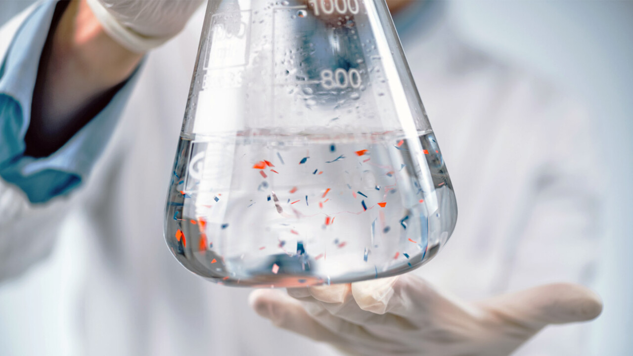 Nahaufnahme eines Laborkolbens mit klarer Flüssigkeit, in der farbige Partikel bzw. Probenbestandteile schweben. Eine Person mit Handschuhen hält den Kolben im Labor – Symbolbild für chemische Analytik, Qualitätskontrolle sowie Untersuchungen zur Lebensmittelsicherheit und Authentizität.