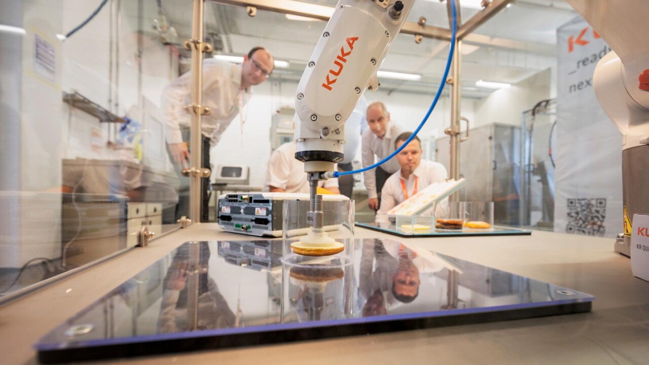 In einem Technikums- bzw. Laborumfeld arbeitet ein KUKA-Industrieroboter an einer Versuchsanlage und greift bzw. positioniert ein Lebensmittelprodukt für Test- oder Prozessschritte. Mehrere Personen beobachten den Ablauf hinter einer Schutzverglasung. Das Bild steht für Automatisierungstechnik, Robotik, digitale Prozessentwicklung und Qualitätssicherung in der modernen Lebensmittelverarbeitung.