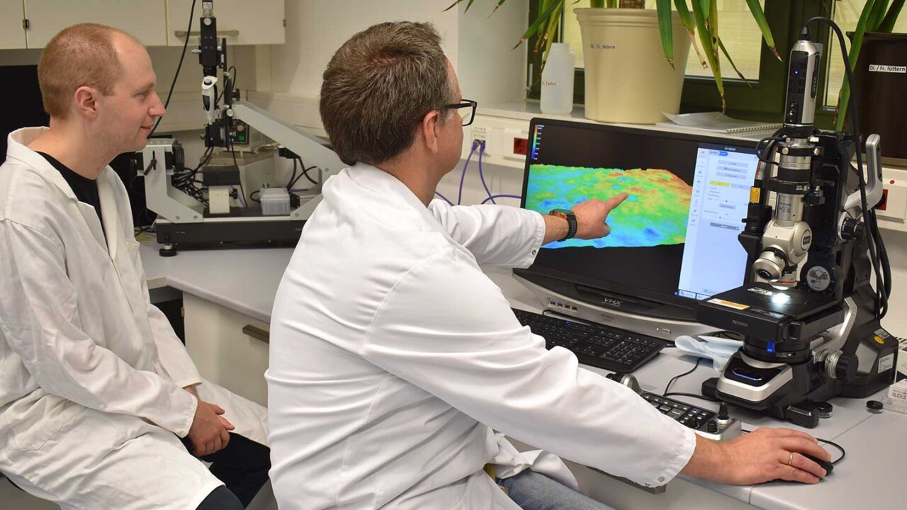 Zwei Wissenschaftler im Laborkittel werten am Monitor eine farbcodierte Oberflächen- bzw. Höhenkarte einer Probe aus. Neben dem Bildschirm steht ein Digitalmikroskop zur hochauflösenden Bildaufnahme. Das Foto steht für Strukturanalyse, Mikrostruktur-Charakterisierung, Struktur-Funktionalitätsforschung und die Bewertung von Verarbeitungseinflüssen auf Produkteigenschaften – z. B. in der Lebensmittel- und Materialforschung.