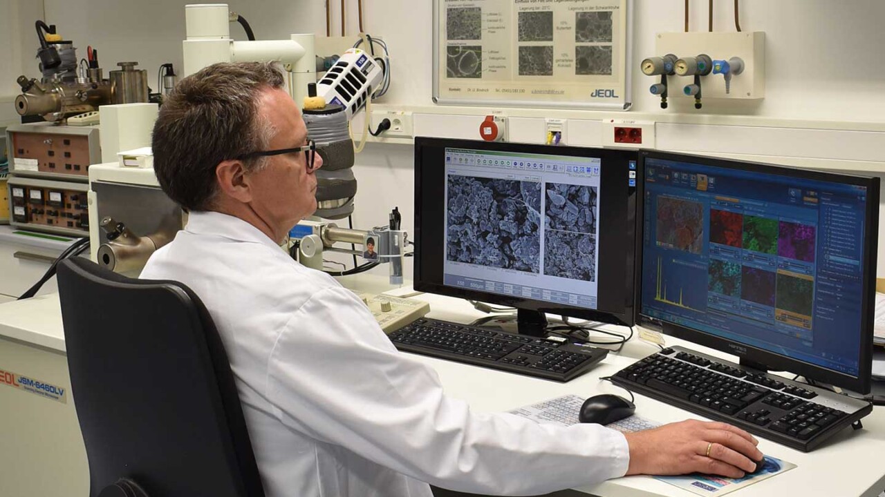 Ein Wissenschaftler im Laborkittel sitzt an einem Auswertungsplatz und analysiert Mikroskopieaufnahmen auf zwei Monitoren. Im Hintergrund ist ein Rasterelektronenmikroskop (REM) zu sehen; auf den Bildschirmen erscheinen hochauflösende Strukturbilder und Auswertedaten. Das Foto steht für Material- und Lebensmittelforschung, Strukturanalyse, Struktur-Funktionalität sowie die Untersuchung von Mikrostrukturen zur Bewertung von Verarbeitung, Qualität und Produkteigenschaften.