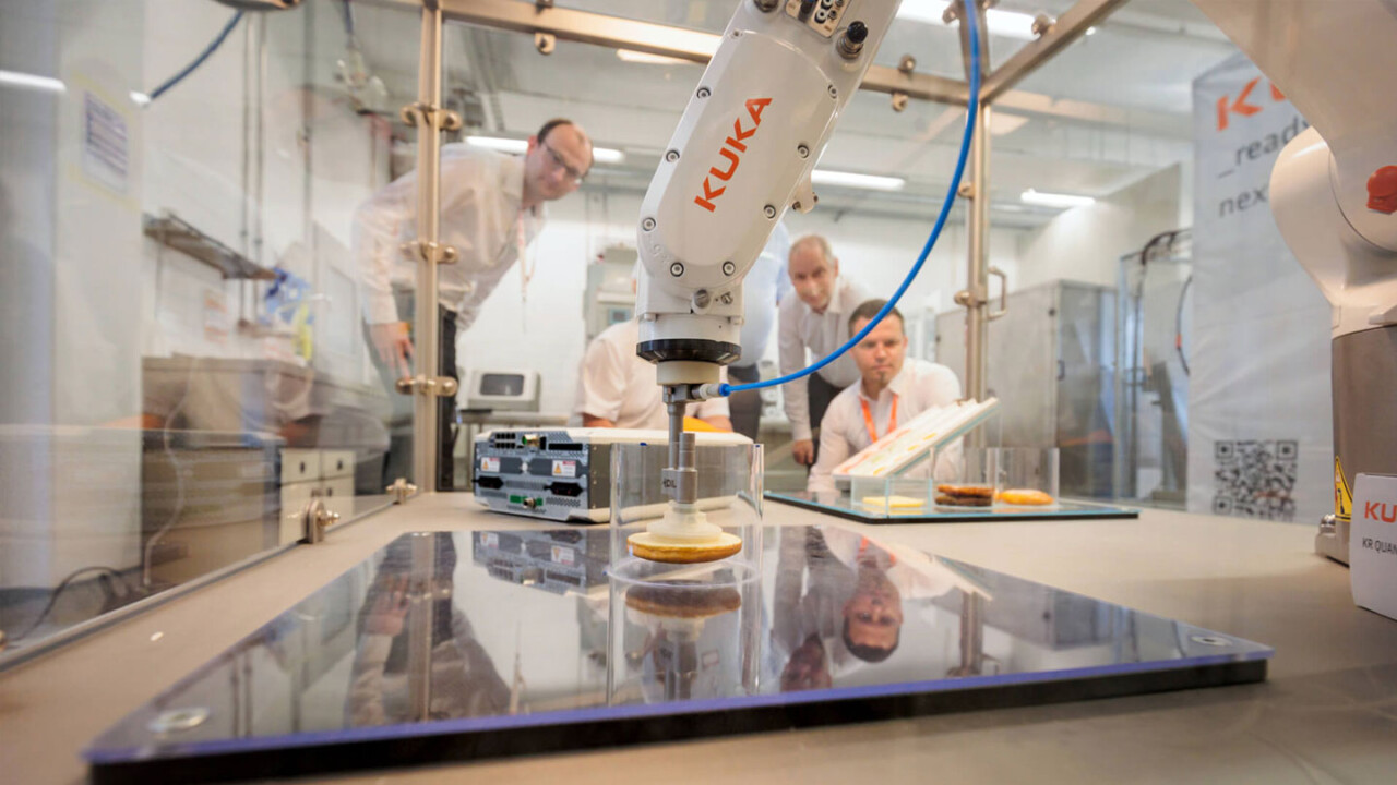 In einem Labor- bzw. Technikumumfeld arbeitet ein KUKA-Roboterarm in einer Sicherheitszelle und greift ein Lebensmittelprodukt auf einer glänzenden Arbeitsfläche. Im Hintergrund beobachten mehrere Personen die Demonstration. Das Motiv steht für Automatisierung, Robotik und innovative Prozessentwicklung in der Lebensmittelproduktion.