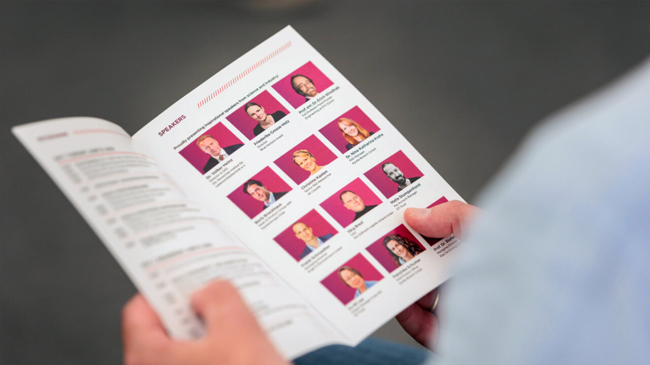 Nahaufnahme einer Person, die ein Veranstaltungsprogramm in den Händen hält und darin blättert. Auf der rechten Seite ist eine „Speakers“-Übersicht mit Porträtfotos und Namen der Referierenden zu sehen. Das Motiv vermittelt die Orientierung im Tagesprogramm und die Vielfalt der Speaker bei einer Fachveranstaltung.
